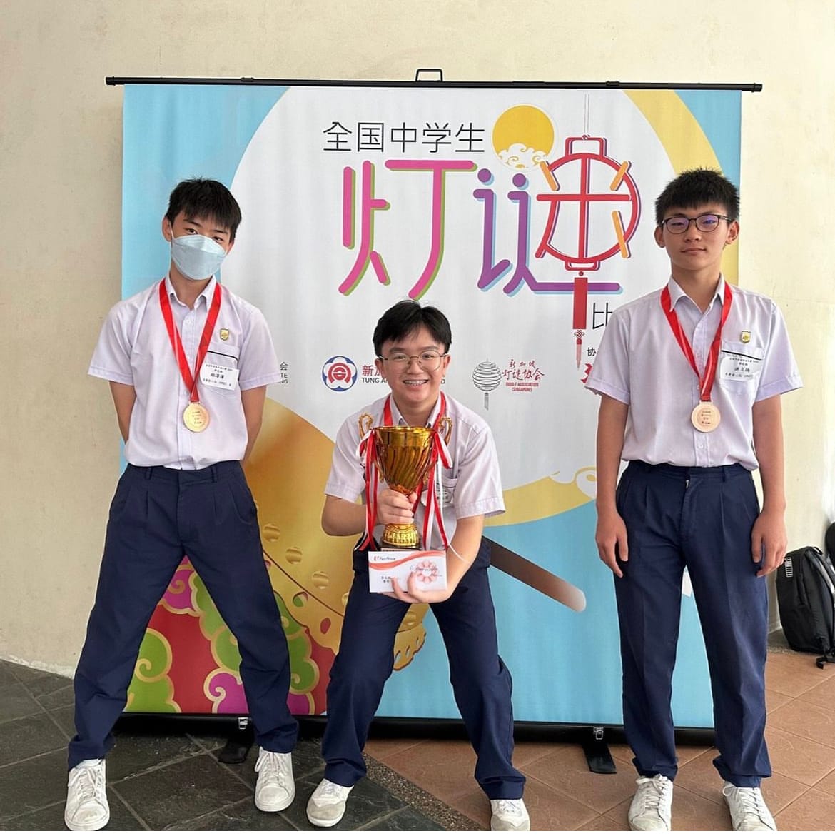 Three male students in school uniforms with medals pose before a Chinese banner; the center boy holds a gold trophy.